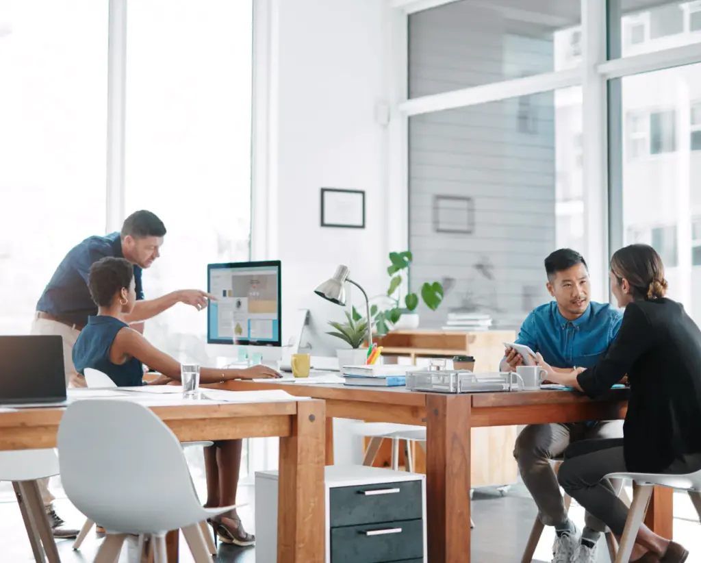 Two pairs of people talking in an office, one pair looking at papers and one at a computer screen